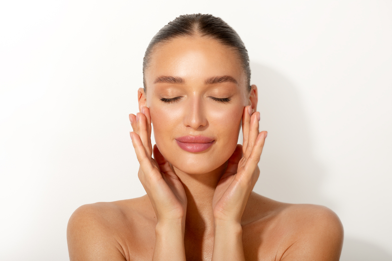 Serene brunette woman with glowing skin cradling her face in hands, eyes closed in relaxation against white background, skincare beauty portrait.