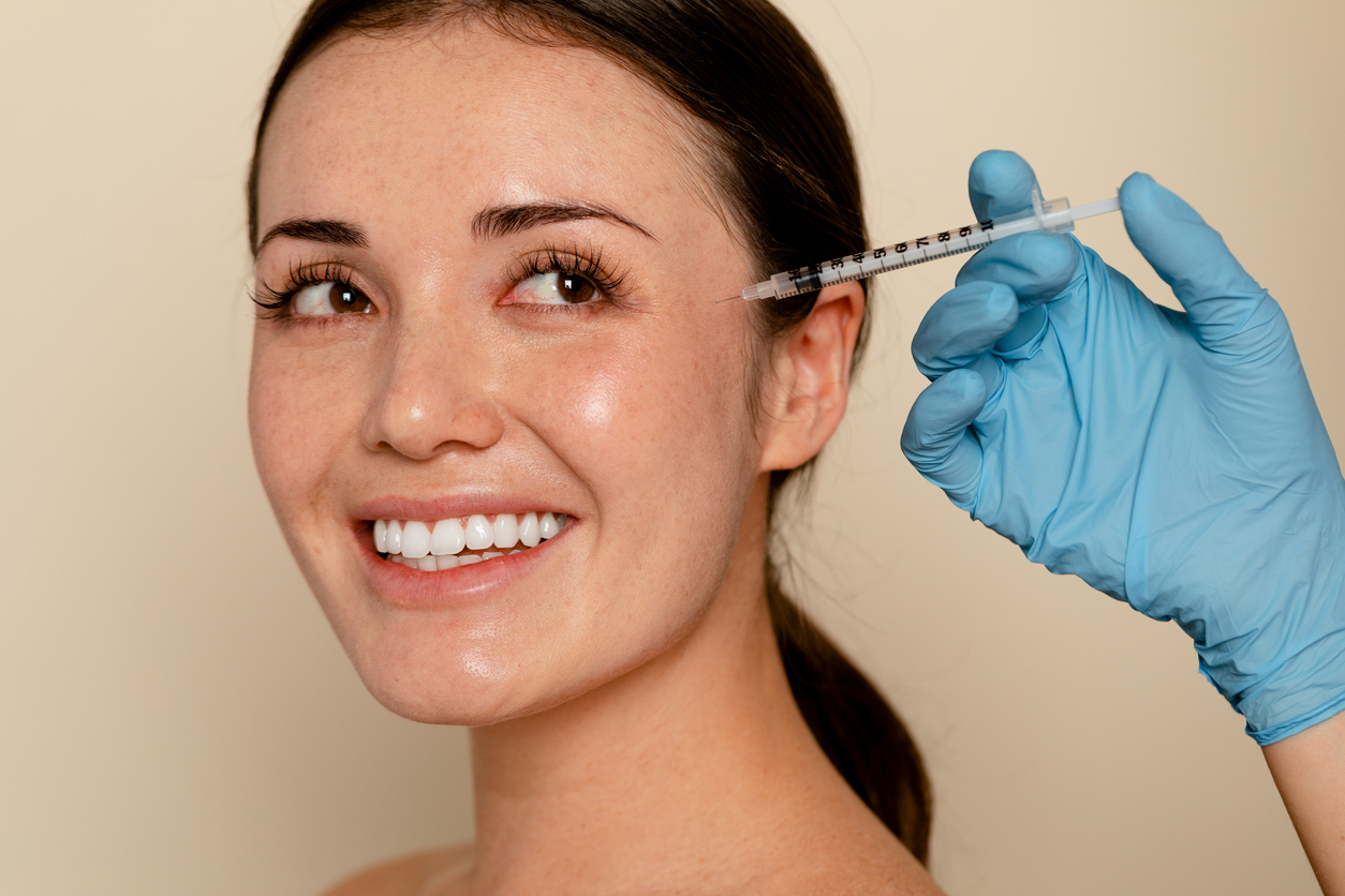 Smiling woman receiving a cosmetic facial injection near the eye area by a medical professional wearing blue gloves, close-up on face against a neutral background.